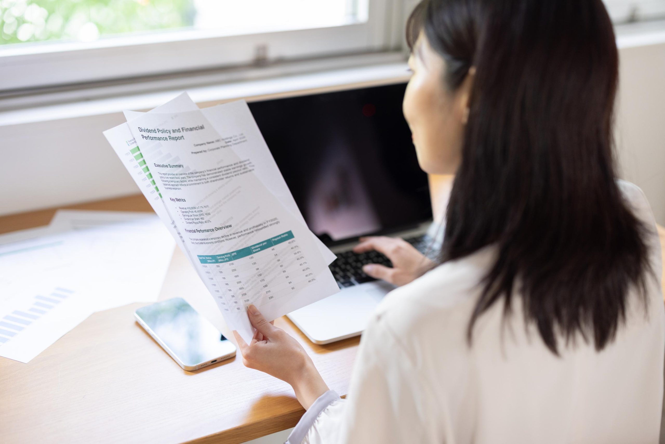 Back view of woman working on laptop while holding document at window
