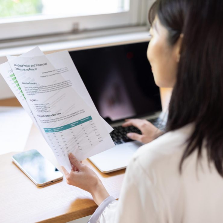 Back view of woman working on laptop while holding document at window