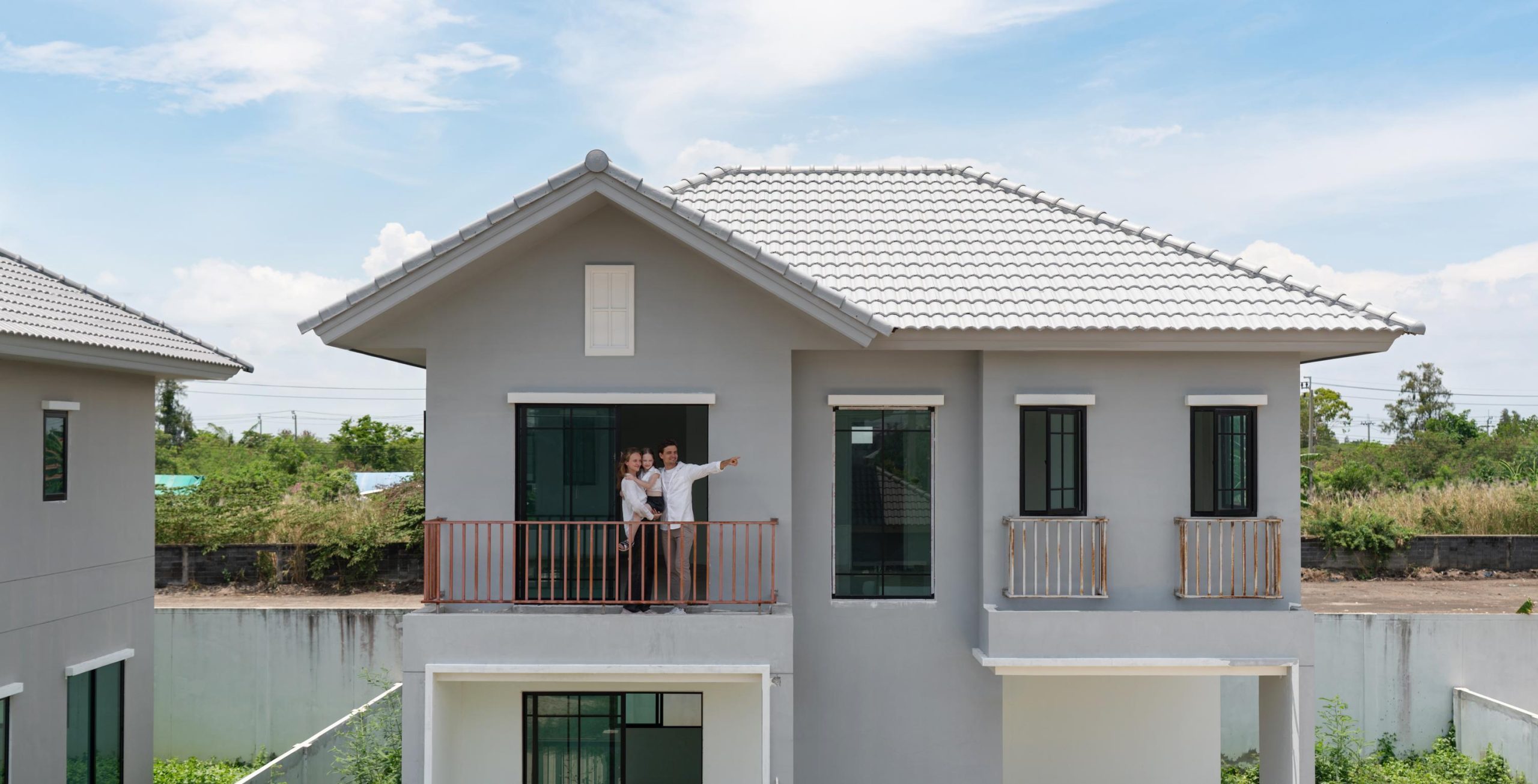 A cheerful family stand on the balcony of a newly constructed modern house, pointing at the landscape. Clear sky and a vibrant setting create a cheerful ambiance in this lively area.
