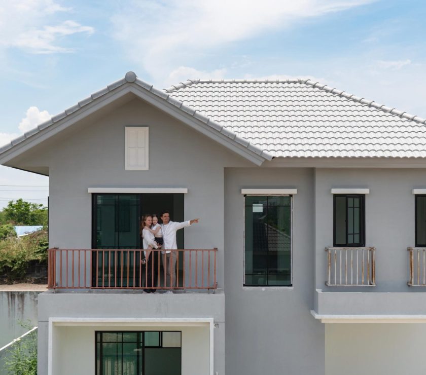 A cheerful family stand on the balcony of a newly constructed modern house, pointing at the landscape. Clear sky and a vibrant setting create a cheerful ambiance in this lively area.