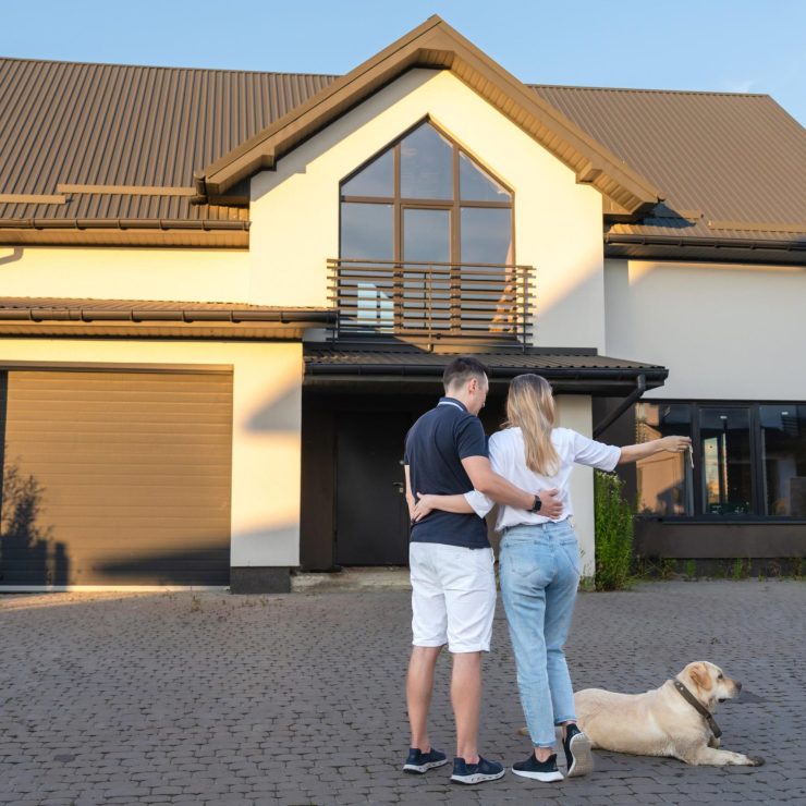 Happy young couple with dog holding their keys in front of residential home. Property lease real estate for relocation. Leasing insurance and purchase. Buying or renting