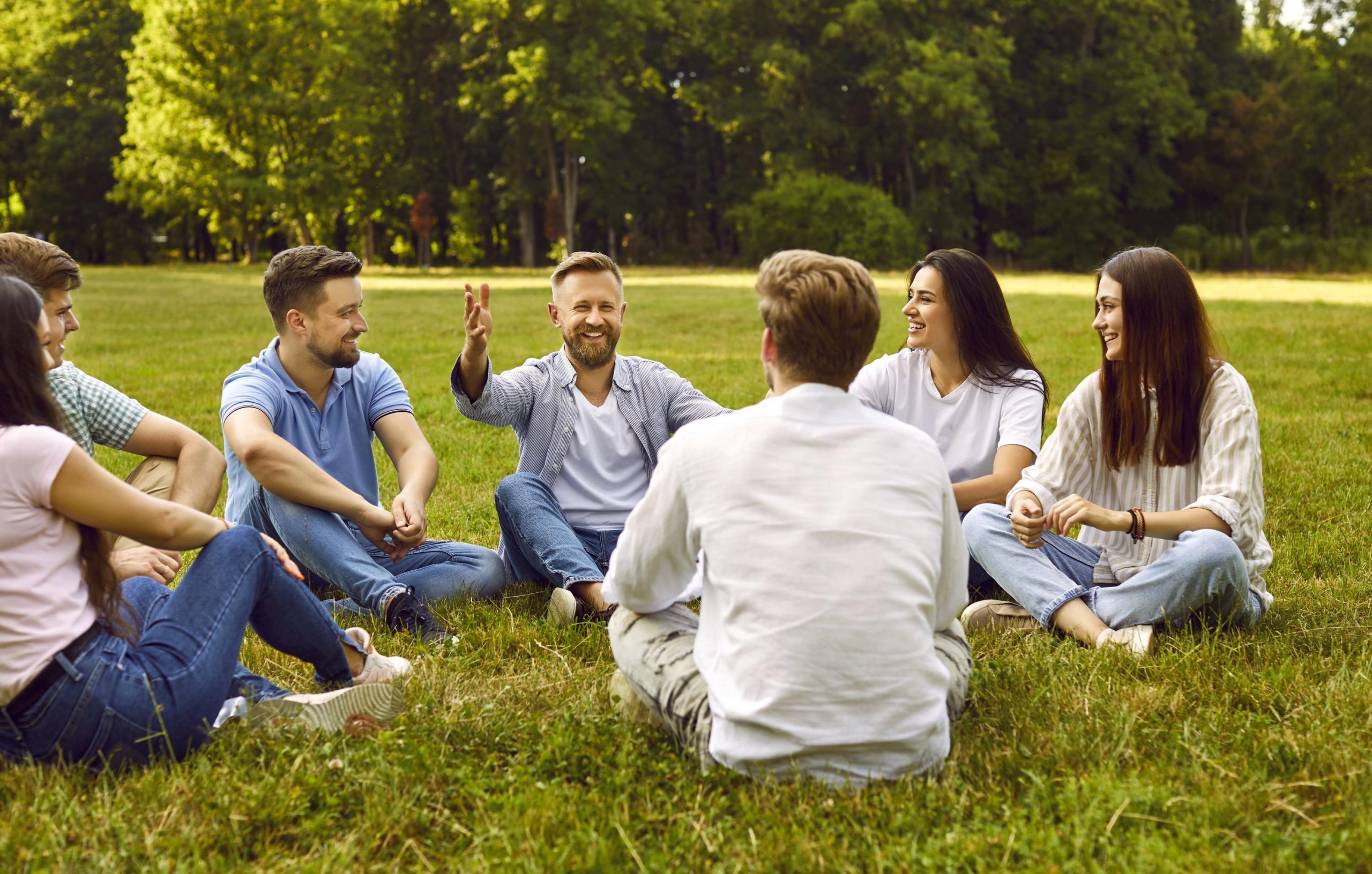 Group of young people communicate sitting on grass in park on summer sunny day.