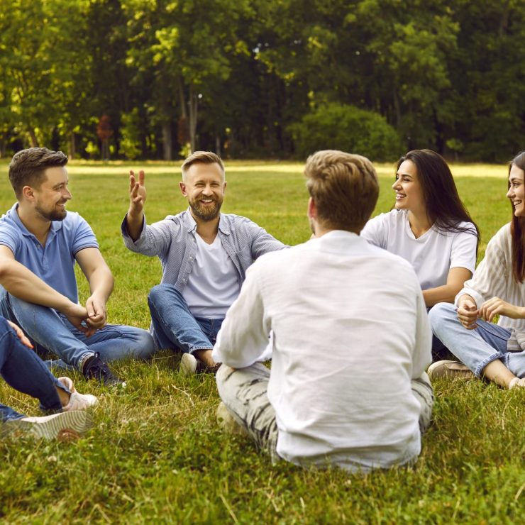 Group of young people communicate sitting on grass in park on summer sunny day.