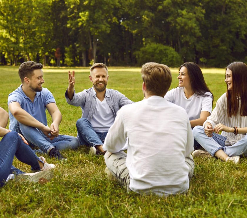 Group of young people communicate sitting on grass in park on summer sunny day.